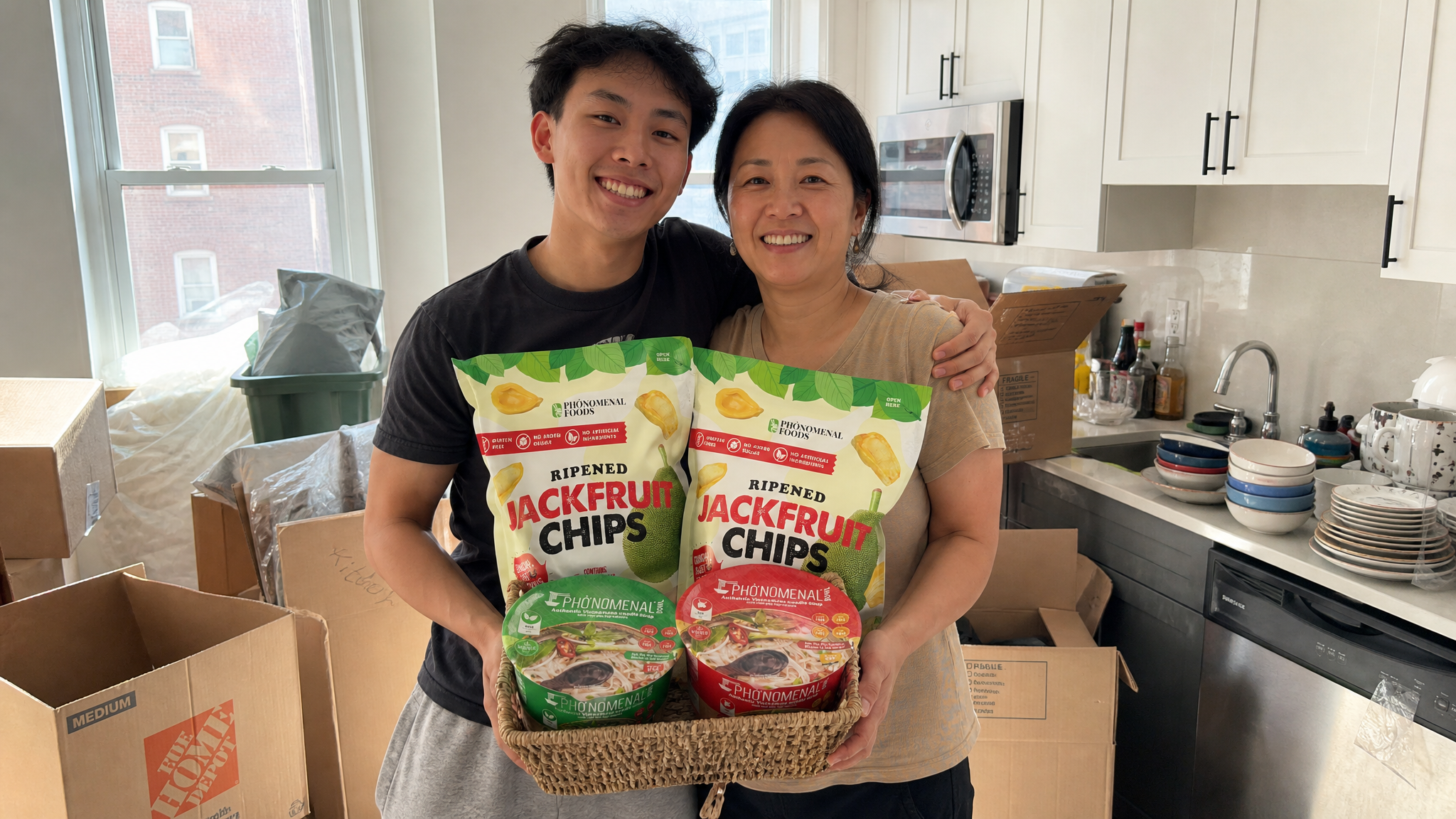 A young man and his mom smiling together in his first apartment kitchen on moving day, holding a gift basket filled with Pho'nomenal Beef and Veggie Pho Bowls and Ripened Jackfruit Chips, with unpacked moving boxes visible in the background.